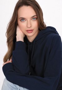 Hanorac bleumarin cu un buzunar frontal, glugă cu șnur și manșete ribbate. Material moale cu o textură netedă, purtat lejer cu blugi deschiși.