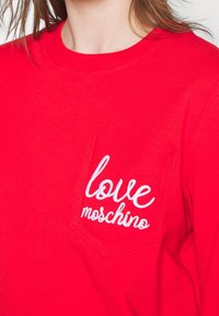 Suéter rojo con cuello redondo, un bolsillo en el pecho izquierdo y texto bordado en blanco que dice "love moschino". Textura de tela suave.