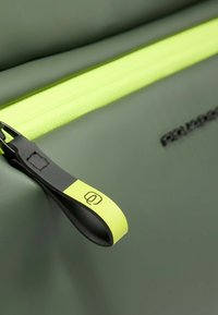 Borsa verde oliva con una cerniera gialla neon brillante. Il tiralampo è nero con un accento giallo e un logo. Materiale liscio e resistente.