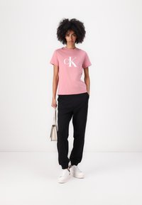 Rosa bomulls T-skjorte med en hvit "CK" logo, kombinert med svarte joggebukser og hvite sneakers. Minimalistisk design med korte ermer.