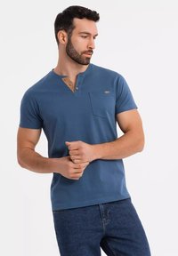 Camisa henley de algodón azul de manga corta con una tapeta de botones color beige, bolsillo en el lado izquierdo del pecho y un diseño ajustado. Se llevan jeans por debajo.
