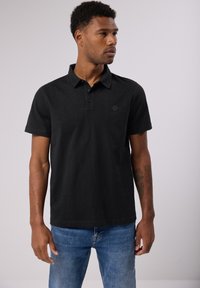 Paita polo musta, valmistettu teksturoidusta kankaasta, jossa on kaulus, kolme nappia ja pyöreä logo vasemmassa rinnassa.