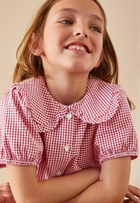Punainen ja valkoinen ruudullinen blusa, jossa on suuri, rypytetty kaulus, lyhyet puff-hihat ja sydämenmuotoiset napit. Silkkinen kankaan rakenne.