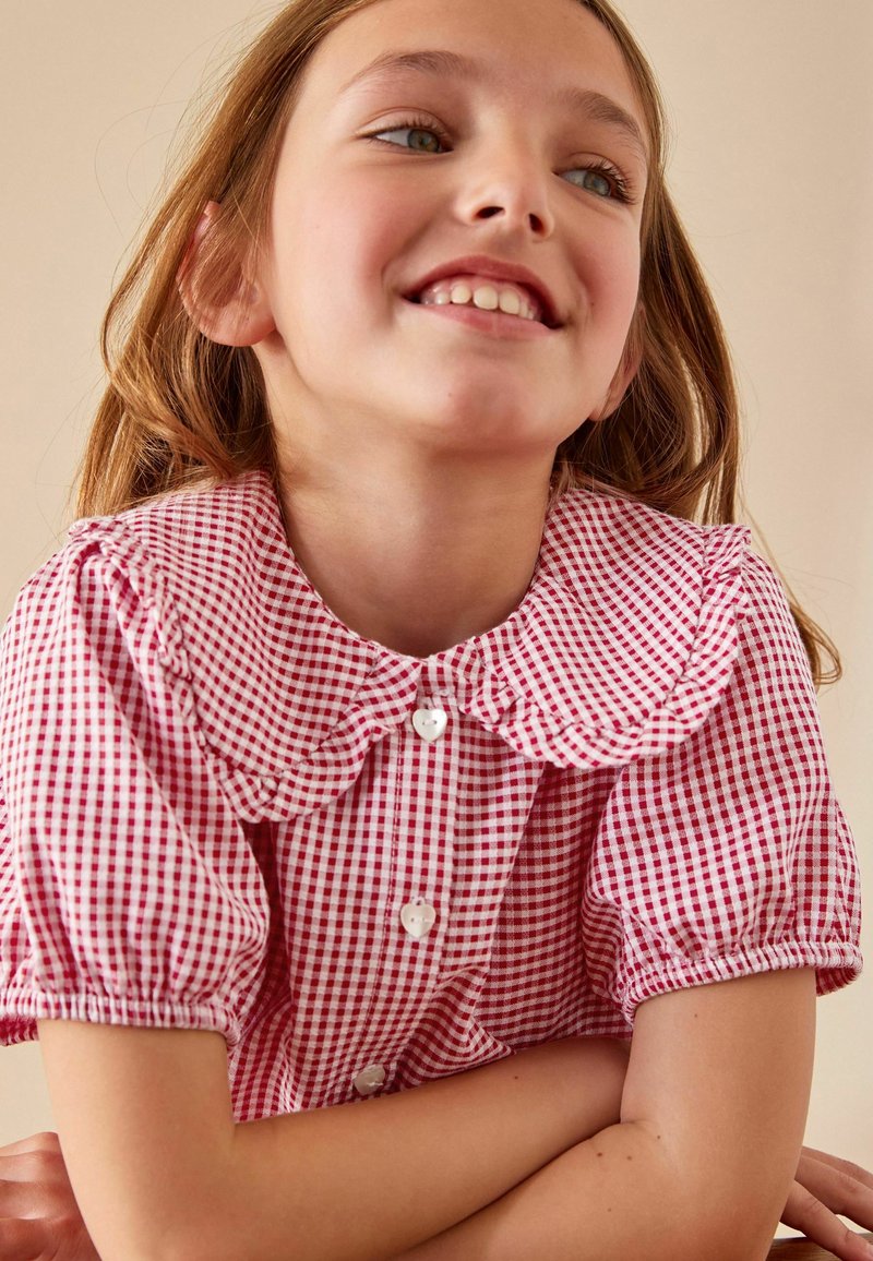Punainen ja valkoinen ruudullinen blusa, jossa on suuri, rypytetty kaulus, lyhyet puff-hihat ja sydämenmuotoiset napit. Silkkinen kankaan rakenne.