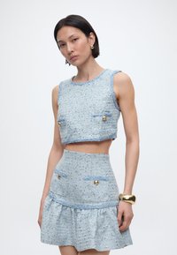 Γυναίκα που φοράει αμάνικο γαλάζιο τοπ με υφή και ταιριαστό κοντό φούστα με ψηλή μέση, διακοσμημένα με χρυσά κουμπιά και χρυσό βραχιόλι μανσέτα.