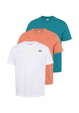 Mand iført hvid kortærmet t-shirt med logo, mørke cargobukser og hvide sneakers, stående med hænderne bag ryggen og kigger mod venstre.