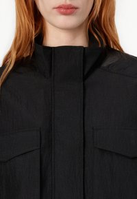 Melnas apkakles jakas ar teksturētu audumu, kurai ir divas krūšu kabatas, tīrs priekšējais dizains un augsta kakla izgriezums.