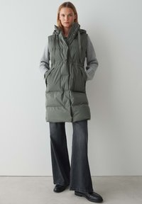 Grüner gepolsterter Weste mit Kapuze, tailliertem Schnitt und seitlichen Taschen, über einem grauen Pullover getragen. Kombiniert mit weiten, dunklen Jeans und schwarzen Stiefeln.