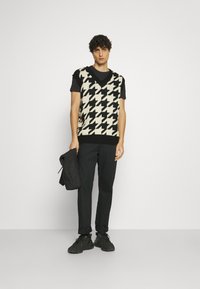 Black and cream houndstooth knit vest over a black t-shirt, paired with black trousers and black sneakers. The model holds a black quilted jacket.