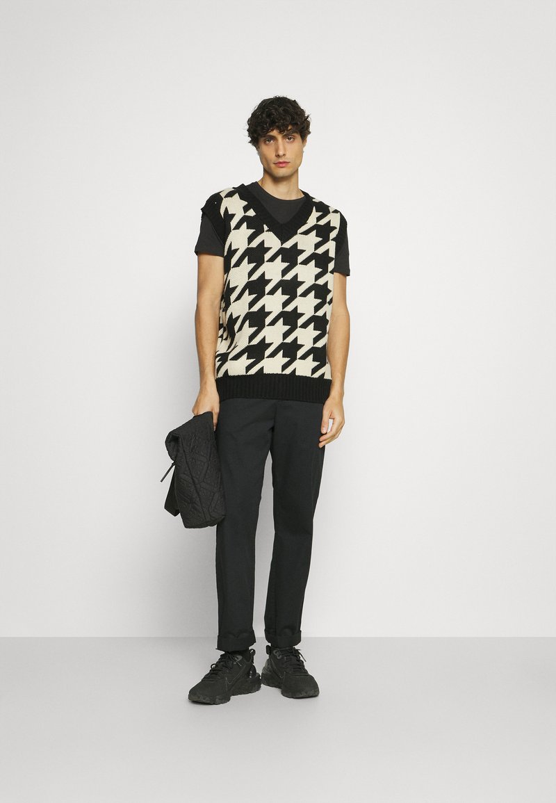 Black and cream houndstooth knit vest over a black t-shirt, paired with black trousers and black sneakers. The model holds a black quilted jacket.