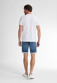 Bílý krátký rukáv T-shirt s malými barevnými vzory, kombinovaný s ohrnutými denimovými šortkami. Model má na sobě bílé tenisky s béžovými akcenty.