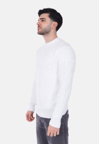 Maglione bianco a maglia cable-knit con collo rotondo e polsini e orlo a coste. La texture è in rilievo con un motivo classico. Indossato con pantaloni grigi.