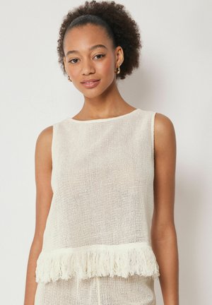 Junge Frau mit lockigem Haar trägt ärmelloses weißes Stricktop mit Fransen am Saum und steht vor einem schlichten, hellen Hintergrund.