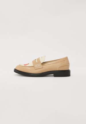 Mocassins en cuir beige avec un dessus blanc contrastant, ornés d'une sangle et d'accents en boucle argentée, posés sur une semelle plate noire.