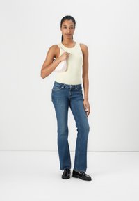 Lys gul tanktop, blå høytalende jeans og svarte loafers. Modellen holder en liten rosa håndveske. Enkle og tettsittende design elementer.
