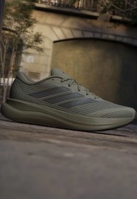 Φωτεινό ελιάς πράσινο παπούτσι δρόμου Adidas με επάνω μέρος από δίχτυ, τρεις ρίγες, κορδόνια και ενδιάμεση σόλα Cloudfoam σε εξωτερικό πεζοδρόμιο.
