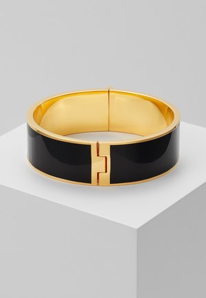 Bracelet rigide à charnière doré et noir reposant sur un socle blanc au fini lisse et brillant.