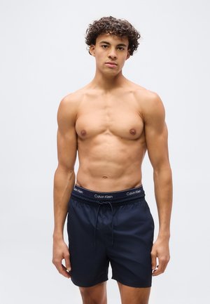 Mladi moški s kodrastimi lasmi, oblečen v temno modre kopalne hlače Calvin Klein, stoji pred enobarvnim svetlo sivim ozadjem.