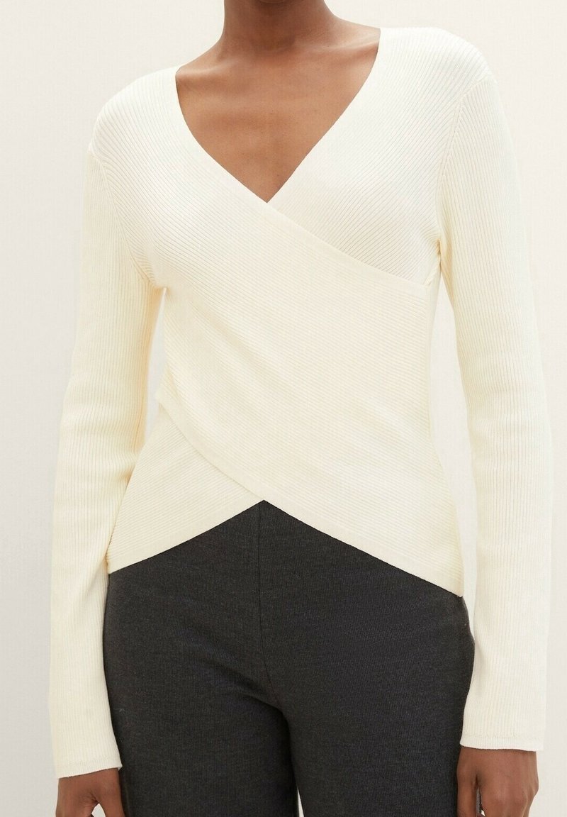 Vit ribbad långärmad topp med djup V-ringning och insvept design, som kombineras med mörkgrå tajta byxor. Slät textur och åtsittande silhuett.