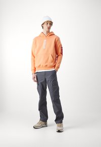 Sudadera naranja con bolsillo frontal, etiqueta "ALPHA", pantalones cargo grises con bolsillos laterales y zapatillas beige con detalles negros. Gorro blanco puesto.