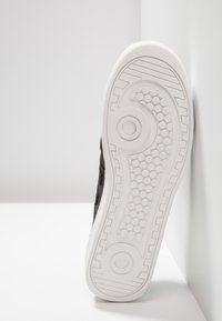 Semelle de basket noire avec une semelle extérieure en caoutchouc blanc. Présente un motif texturé avec des lignes circulaires et horizontales pour l'adhérence.