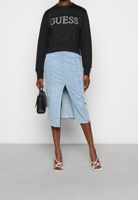 Personne portant un sweat à capuche noir avec le logo "GUESS", une jupe midi en denim bleu clair avec une fente sur le devant, des talons blancs à lanières, tenant un sac à main noir.