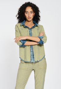 Light green denim shirt with a dark blue collar and cuffs, featuring two front pockets and a relaxed fit. Worn with matching pants.