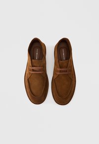 Καφέ σουέτ παπούτσια με στρογγυλεμένο σχήμα, που διαθέτουν δύο καφέ κορδόνια και αντίθετη ραφή κατά μήκος των άκρων. Απλός σχεδιασμός και ομαλή υφή.