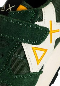 Primo piano di una sneaker in suede verde scuro con un triangolo giallo ricamato e una croce su una toppa di pelle bianca.