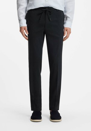 Mężczyzna w czarnych spodniach na sznurki, jasno-niebieskiej koszuli w paski i czarnych butach z beżową podeszwą, stojący na białym tle.