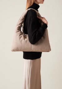 Sac à main texturé beige avec une forme unique, doté d'un design doux et rembourré ainsi que d'une poignée élégante. Porté contre un pull noir.