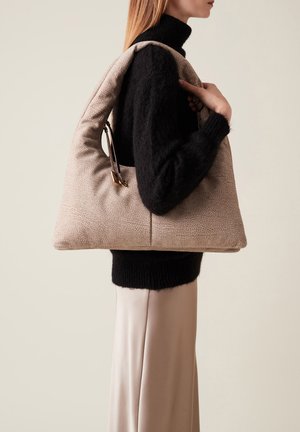 Sac à main texturé beige avec une forme unique, doté d'un design doux et rembourré ainsi que d'une poignée élégante. Porté contre un pull noir.