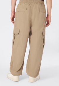 Beżowe spodnie cargo z elastycznym pasem i wieloma kieszeniami na klapki, noszone z białymi sneakersami, widoczne z tyłu na osobie na jednolitym tle.
