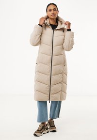 Licht beige gewatteerde jas met een capuchon, voorzien van een voorkant met rits, een chevronpatroon en twee zijzakken. Gekleed met denim en sneakers.