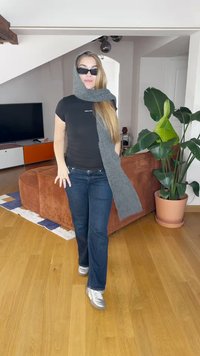 Black fitted t-shirt, dark blue jeans, grey scarf, and silver athletic shoes. Indoor setting with a brown sofa and green plant.
