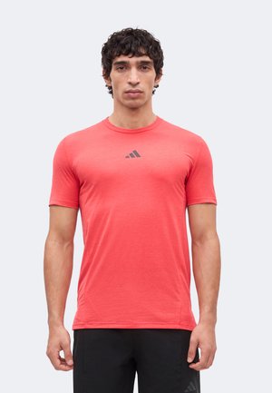 Mann trägt korallenfarbenes, eng anliegendes Adidas-T-Shirt mit kurzen Ärmeln und schwarze Adidas-Hose, steht vor einfarbigem hellgrauen Hintergrund.