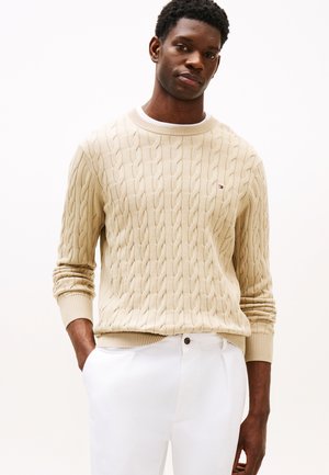 Beige kabelstickad tröja med ribbade muddar och fåll, med subtila horisontella linjer. Bärs med vita byxor och visas mot en neutral bakgrund.
