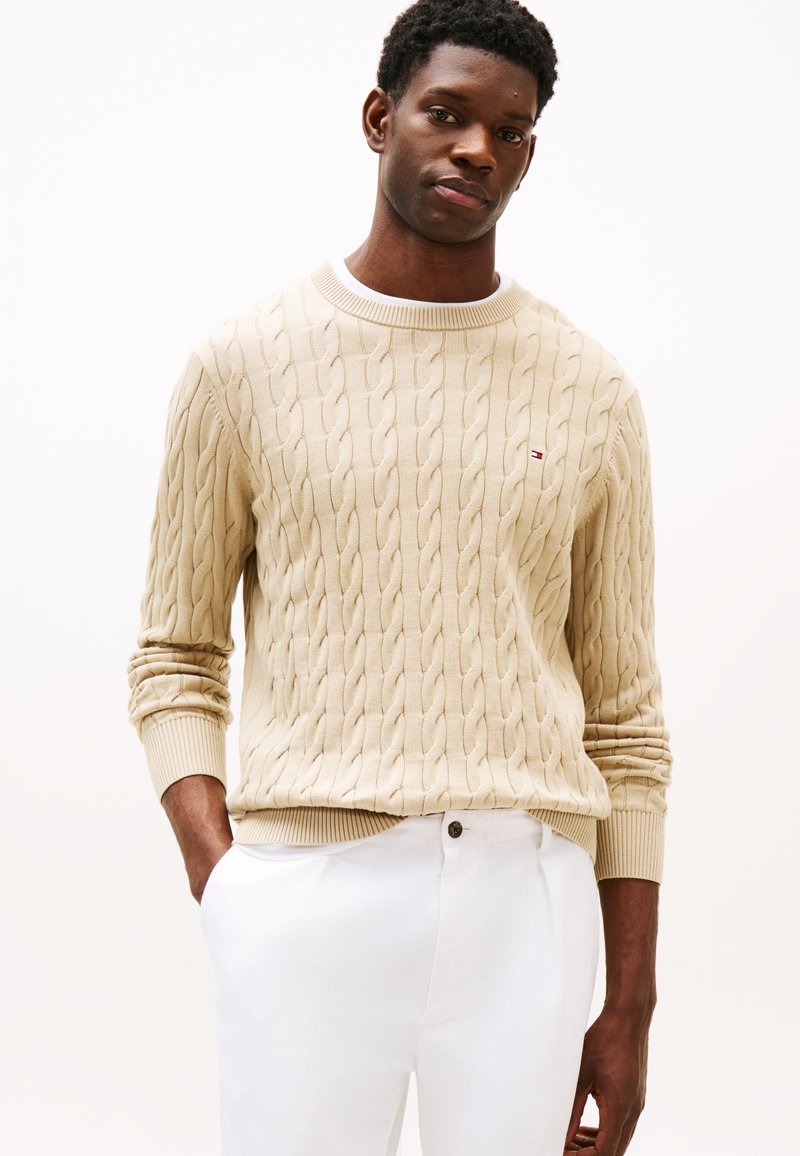 Μπεζ πουλόβερ με πλεκτή υφή και κορδόνια στις μανσέτες και το τελείωμα, με διακριτικές οριζόντιες γραμμές. Φορεμένο με λευκό παντελόνι, παρουσιάζεται σε ουδέτερο φόντο.