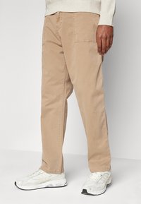 Beige Baumwollhosen mit lockerer Passform, ausgestattet mit einer Seitentasche und sichtbaren Nähten. Kombiniert mit weißen Sportschuhen.
