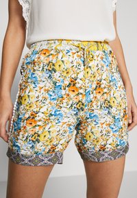Květinové šortky na bílém pozadí, zdobené žlutými, oranžovými a modrými květy. Elastický pas se žlutou šňůrkou a vzorovaným lemem.