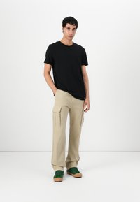 Tricou negru din bumbac, pantaloni cargo bej cu buzunare laterale și adidași verzi cu tălpi maro, stând pe un fundal alb.
