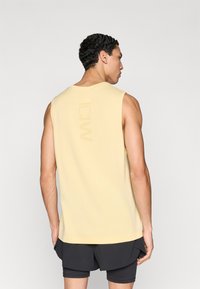 Αμάνικη κίτρινη φανέλα από απαλό ύφασμα, με στρογγυλή λαιμόκοψη και ένα διακριτικό λογότυπο σε ελαφρώς πιο σκούρα απόχρωση στην πλάτη.