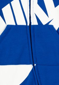 Hoodie azul de algodão com fecho éclair, apresentando um grande design gráfico branco, bolsos canguru e costuras visíveis ao longo dos fechos e costuras.