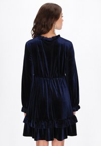 Tengerészkék bársony ruha hosszú ujjakkal, fodros nyakvonallal, derékban összébb húzva, réteges fodros szegéllyel. Sima textúra, elegáns kialakítás.