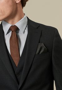 Marineblaues maßgeschneidertes Anzug mit strukturiertem Stoff, hellblauem Hemd, brauner Krawatte und schwarzem gemustertem Taschentuch. Die geknöpfte Weste vervollständigt den Look.