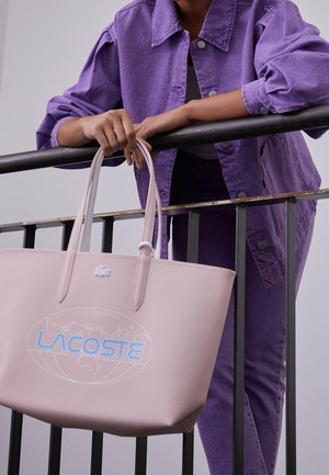 Personne portant une veste et un pantalon en denim violet, tenant un grand sac cabas Lacoste rose clair avec un logo bleu près d'une rambarde noire.