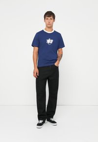 Camiseta de algodón azul marino con cuello blanco y un logo gráfico en el pecho, combinada con vaqueros negros de pierna recta y zapatillas negras.