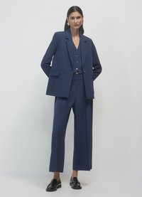 Costume trois pièces sur mesure bleu comprenant un blazer, un gilet et un pantalon à jambes larges, avec un tissu lisse et une silhouette décontractée.