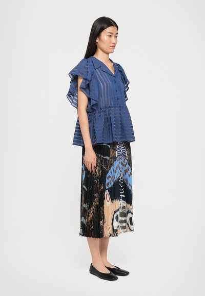 Woman wearing a blue striped blouse with ruffled sleeves and a pleated, multicolor patterned midi skirt, standing in profile with black flats.