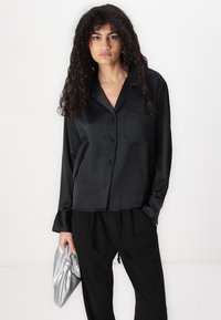 Black satin long-sleeve shirt with a collar, front buttons, and a left chest pocket, paired with black pants and holding a silver clutch.